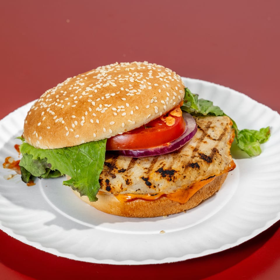 Grilled chicken burger with lettuce, tomato, red onion, sauce, and a sesame seed bun on a white plate.