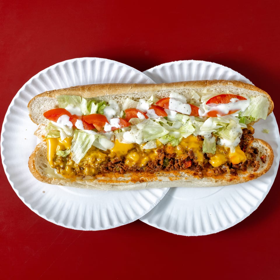 Loaded sub sandwich with seasoned meat, melted cheddar, lettuce, tomatoes, and creamy dressing on a white paper plate against a red background.