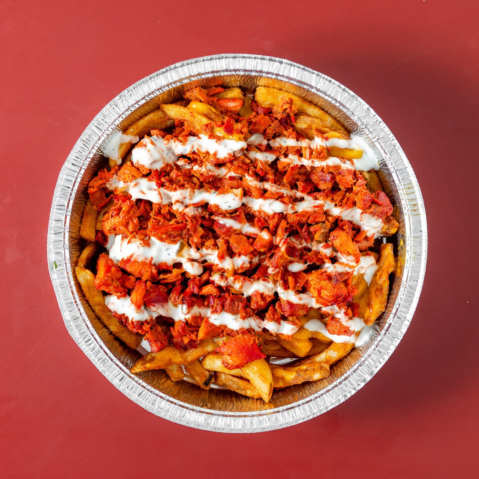 Loaded fries topped with seasoned shredded meat and creamy white sauce in a foil tray on a red background.