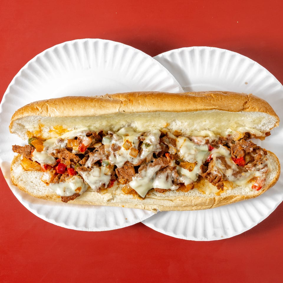Cheesesteak sandwich with melted cheese, sliced steak, peppers, and onions on a long roll atop white paper plates.