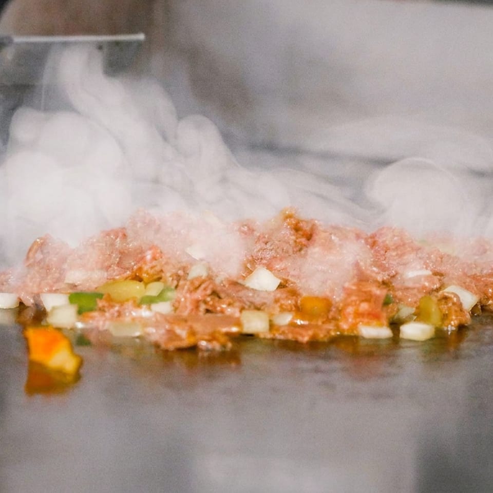 Seasoned ground meat and chopped vegetables sizzle on a flat-top grill, surrounded by rising steam.
