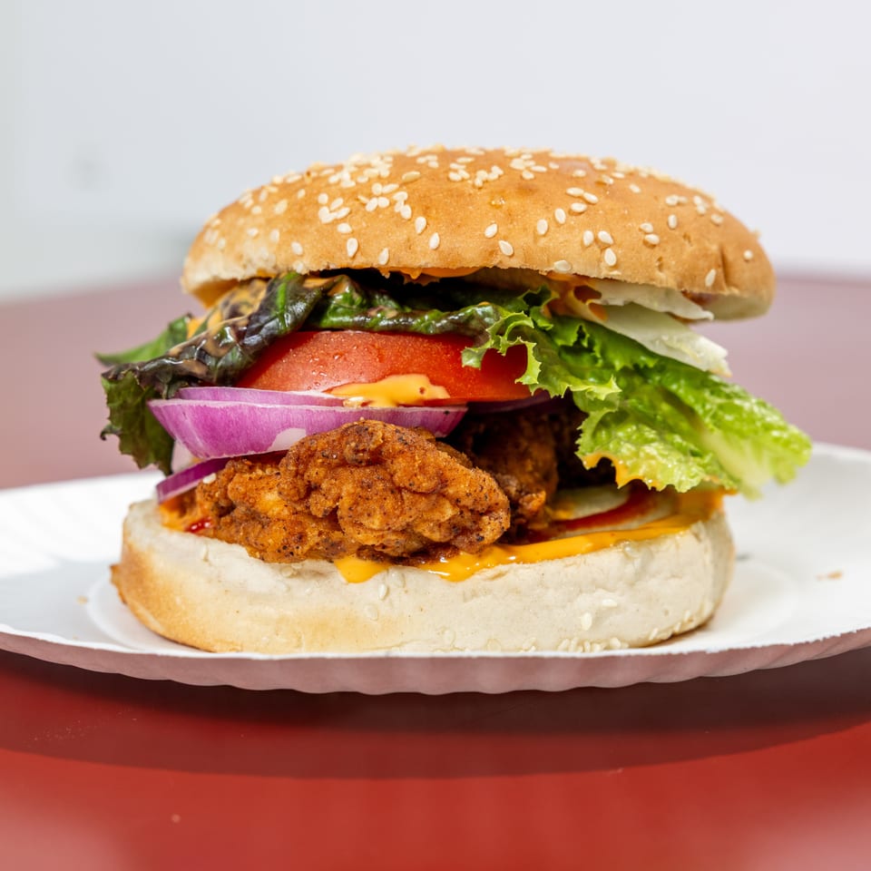Crispy fried chicken burger on a sesame bun with lettuce, tomato, red onion, and cheese sauce on a white plate.