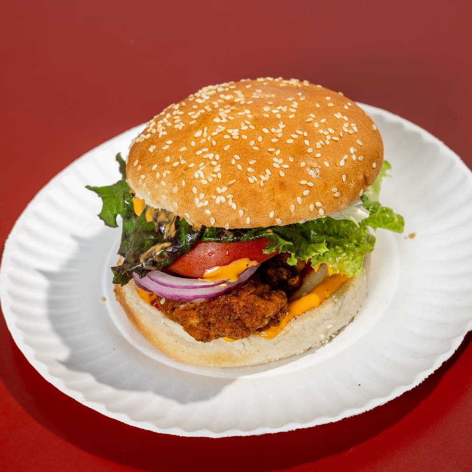 Crispy fried chicken burger with sesame bun, lettuce, tomato, red onion, cheese, and sauce on a white plate.