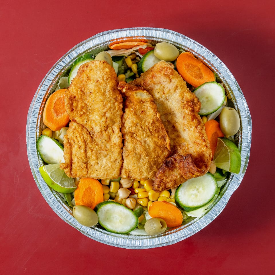 Top-down view of a foil bowl filled with crispy fried chicken over lettuce, cucumber, carrots, corn, olives, chickpeas, and lime.