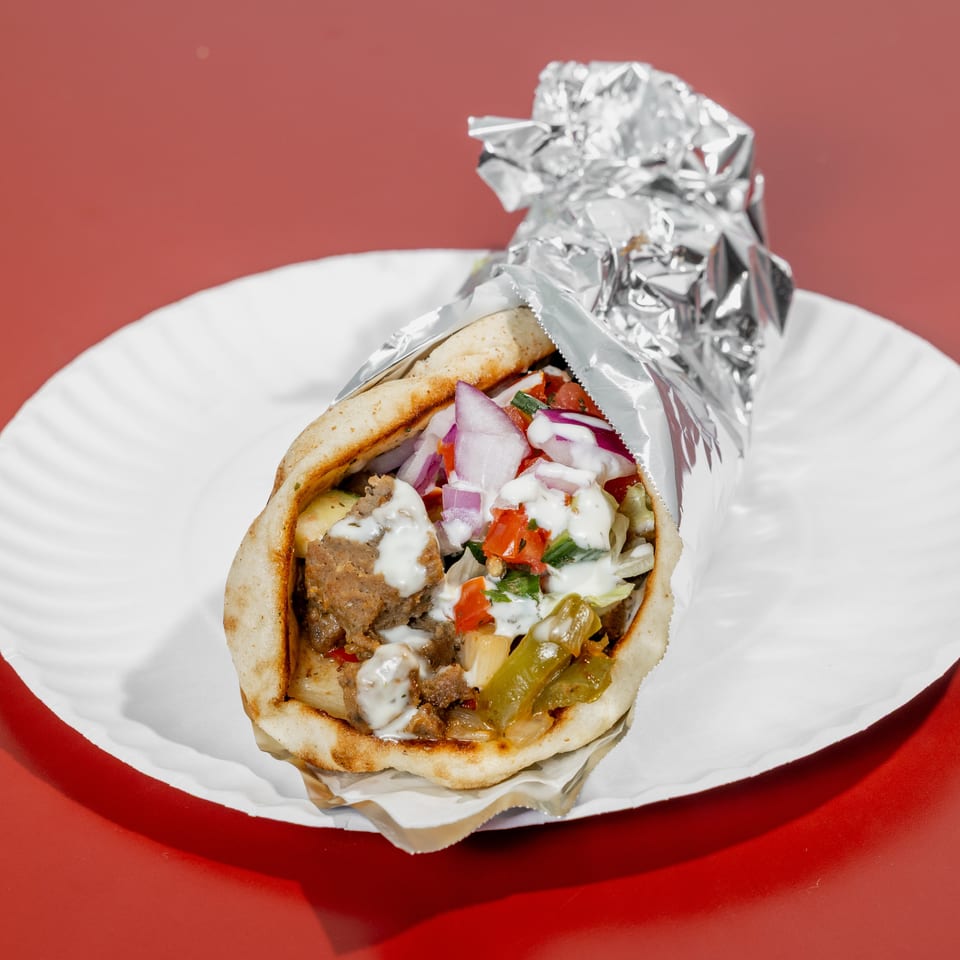 Gyro pita stuffed with seasoned meat, onions, tomatoes, peppers, lettuce, and white sauce on a paper plate.