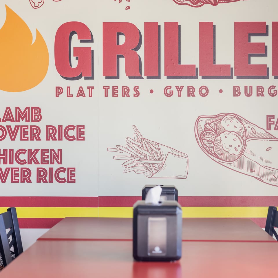 Restaurant dining area with red tables, black chairs, and a colorful mural advertising grilled platters, gyros, burgers, and rice dishes.