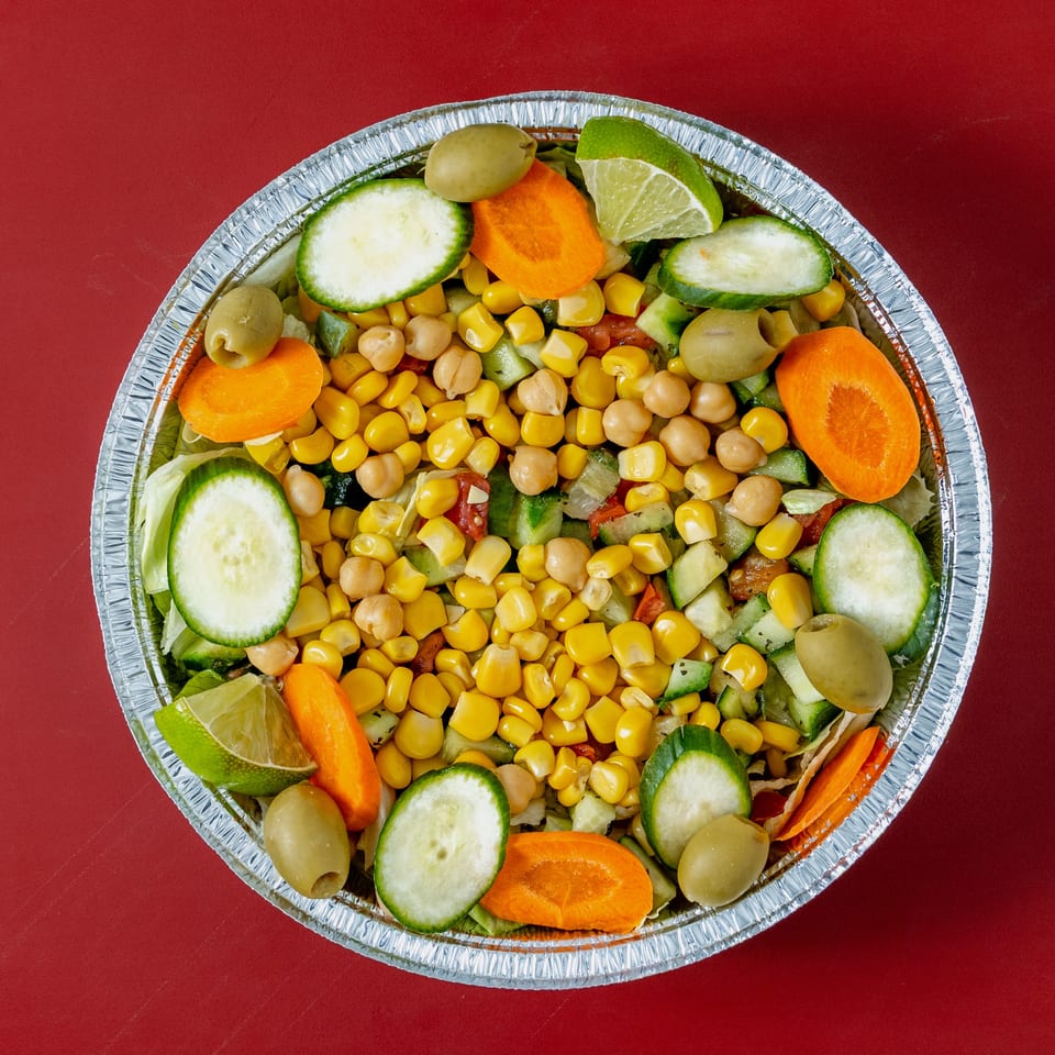 Overhead view of a colorful salad with corn, chickpeas, cucumbers, carrots, olives, lime, and lettuce in a foil bowl.