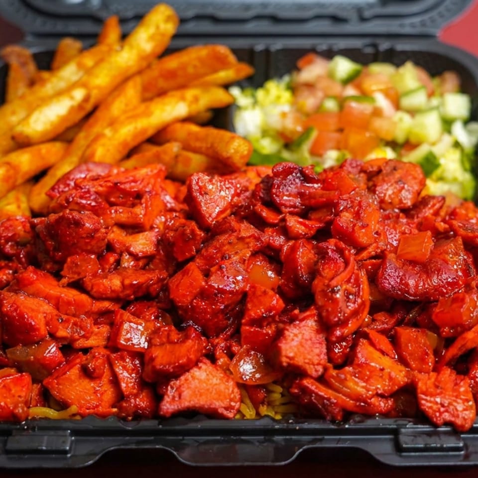 Black takeout tray filled with red-spiced grilled meat and vegetables, seasoned fries, and chopped salad on a red background.