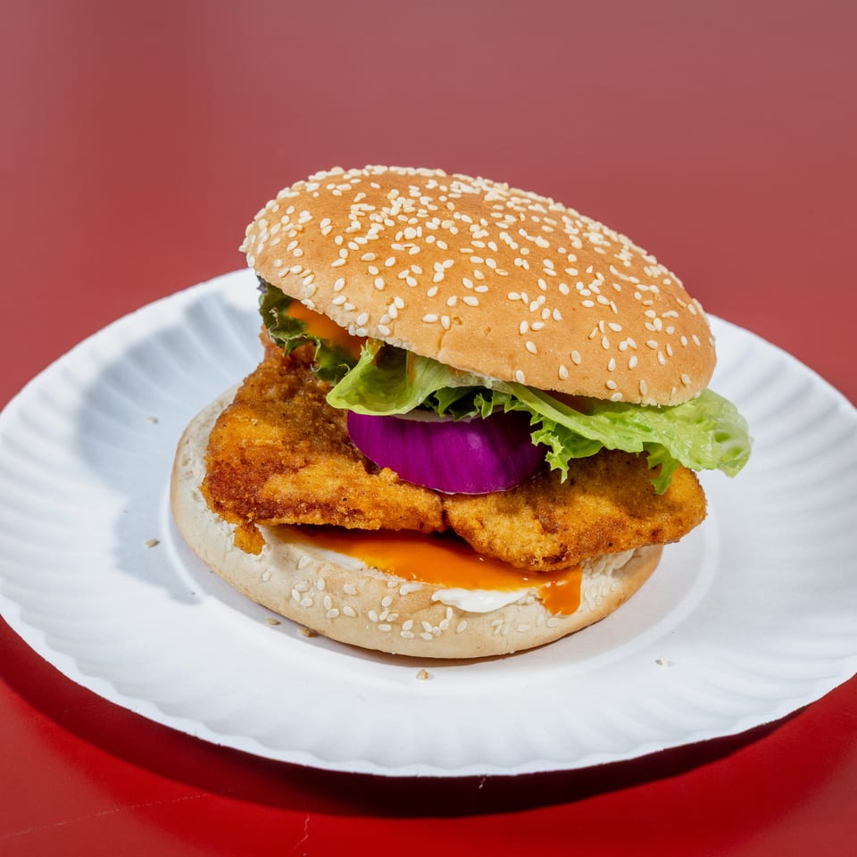 Sesame-seed burger with crispy breaded filling, lettuce, red onion, and sauce on a white plate against a red background.