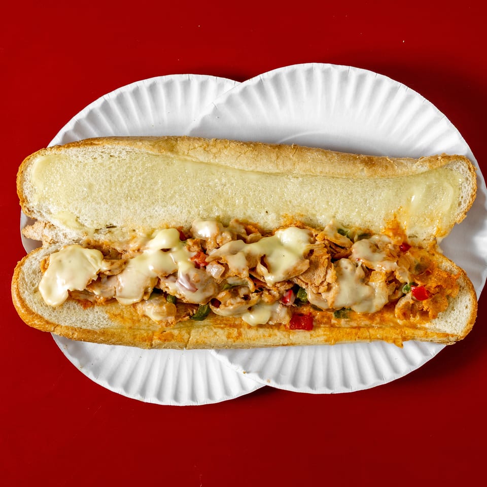 Cheesy chicken sandwich with peppers and onions on a long toasted roll, served on a white paper plate against a red background.