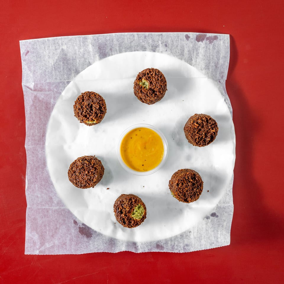 Crispy falafel balls arranged around a cup of yellow dipping sauce on a paper-lined plate against a red background.