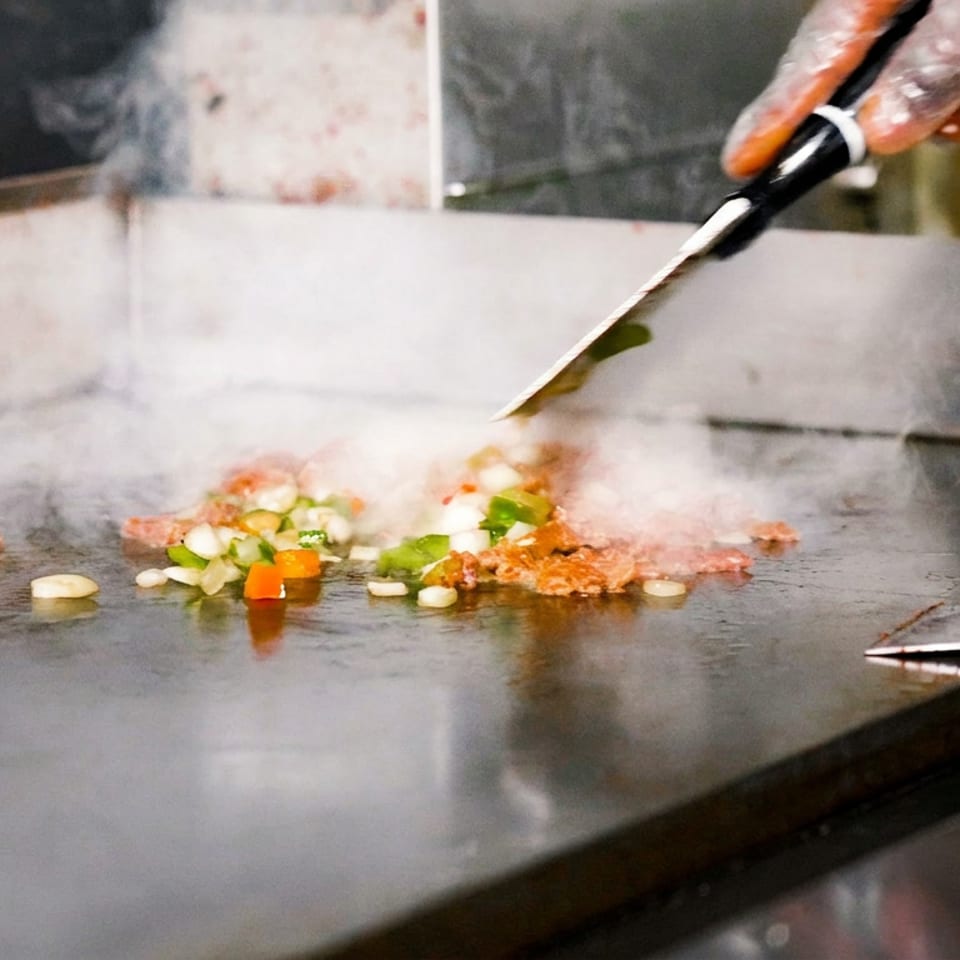 Chef stir-fries chopped vegetables and meat on a steaming flat-top griddle using a spatula.