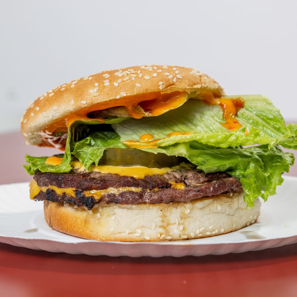 Double cheeseburger with lettuce, pickles, orange sauce, and a sesame-seed bun on a white plate.